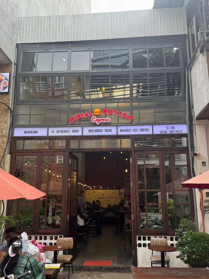 Street view of Bikini Bottom Express in Da Nang, Vietnam — photographed by Mike & Stacy Abroad for Eat · Stay · Play. The open café entrance, red umbrellas, and warm lighting highlight its friendly breakfast atmosphere and popular expat appeal.