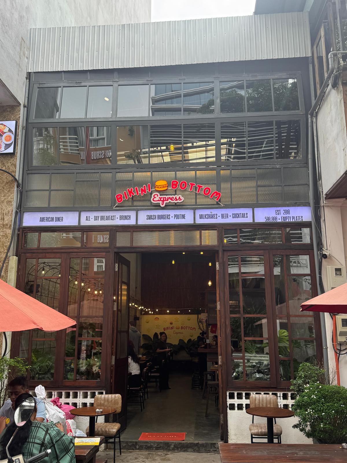 Street view of Bikini Bottom Express in Da Nang, Vietnam — photographed by Mike & Stacy Abroad for Eat · Stay · Play. The open café entrance, red umbrellas, and warm lighting highlight its friendly breakfast atmosphere and popular expat appeal.