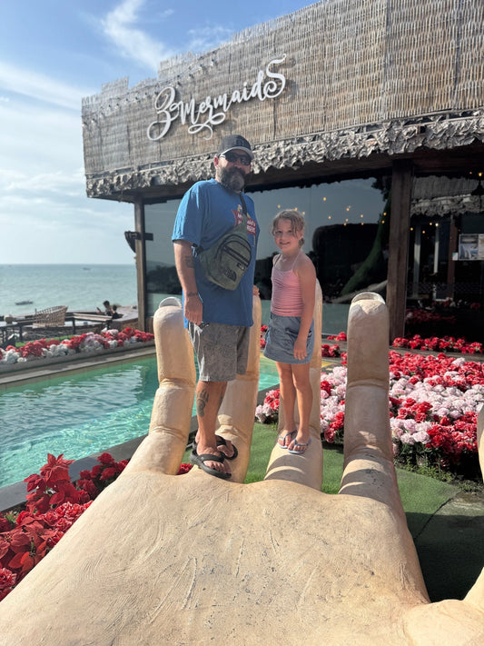 Mike and Hadley standing on a giant hand sculpture at 3 Mermaids Pattaya, with the ocean and café sign behind them — a sunny moment capturing family travel and coastal views in Thailand.