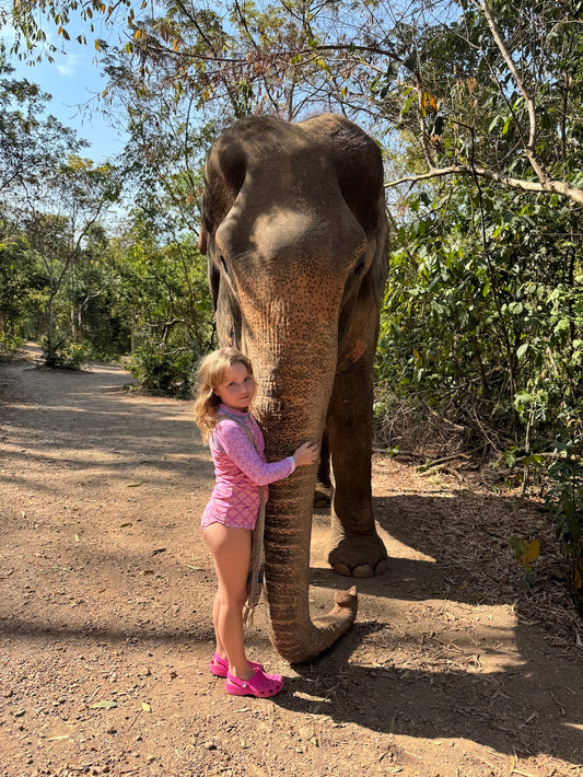 A young girl in a pink long-sleeve swimsuit stands gently hugging an Asian elephant’s trunk in a shaded forest trail at the Pattaya Elephant Sanctuary. Sunlight filters through the trees as she connects calmly with the elephant, capturing a quiet, meaning