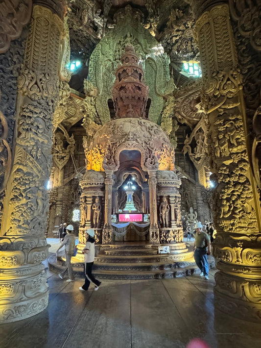 The interior of the Sanctuary of Truth in Pattaya, showing towering carved wooden pillars and an ornate central shrine, with visitors in hard hats walking through the intricate hall.