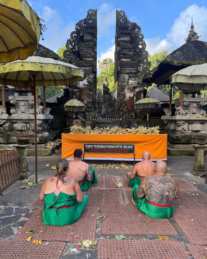 Visitors dressed in traditional green sarongs sit cross-legged on temple mats at the Tirta Empul Water Temple in Ubud, Bali, during a moment of prayer before the water purification ritual. Offerings of flowers and fruit rest on an altar beneath ornate sto