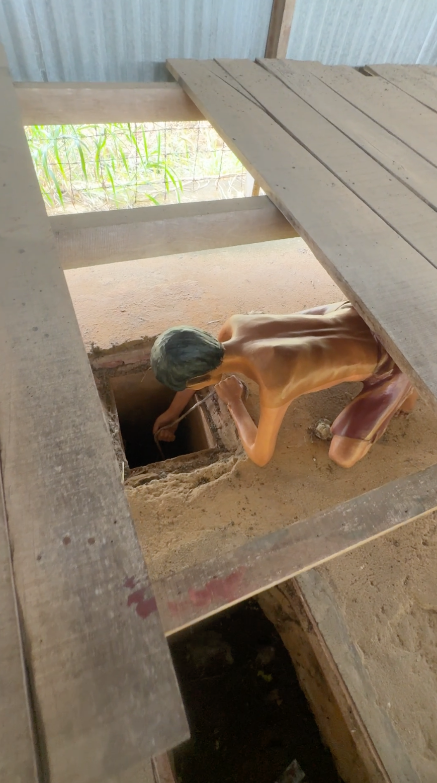 A life-sized figure of an emaciated Vietnamese prisoner kneeling beside a narrow underground escape tunnel inside Phú Quốc Prison, showing how inmates secretly dug through the dirt floor beneath wooden planks in desperate attempts to flee captivity.