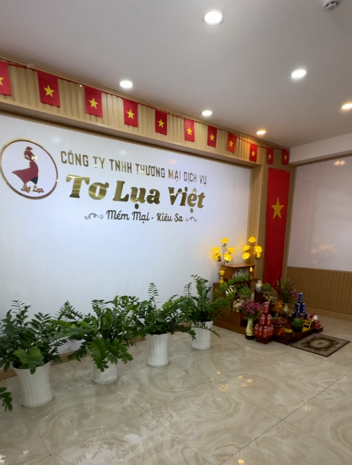 A bright interior view of Tơ Lụa Việt silk shop in Phu Quoc, featuring a white and gold wall with the store’s logo, a row of lush green plants in white pots, and a traditional Vietnamese offering altar decorated with flowers and gifts beneath red flags. A