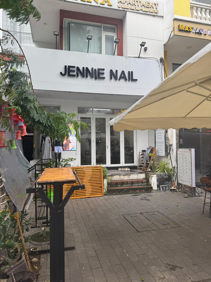 Front entrance of Jennie Nail Spa in Da Nang, Vietnam — a simple white storefront with bold black lettering, steps leading up to the glass doors, and a quiet courtyard space out front on an overcast day. Captured for our Jenny’s Nail & Spa Da Nang review 